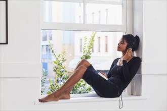 Mixed Race woman sitting in windowsill listening to headphones
