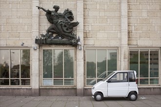 Compact car parked under sculpture on building