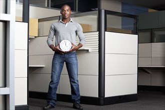 Mixed race businessman holding clock in office cubicle