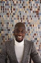 African man in tweed jacket smiling in front of tiled wall