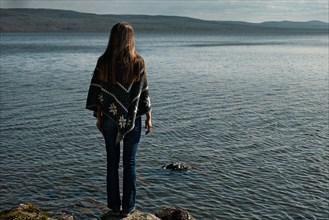 Pensive Caucasian girl standing on rock at lake