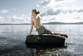Topless Caucasian teenage girl sitting on rock in ocean