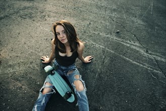 Caucasian teenage girl sitting on pavement with skateboard