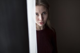 Serious Caucasian woman standing near curtain at window