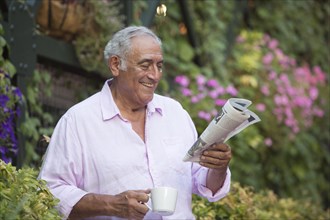 Hispanic man drinking coffee and reading newspaper in flower garden