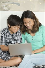 Smiling Asian mother and son sitting on bench using laptop