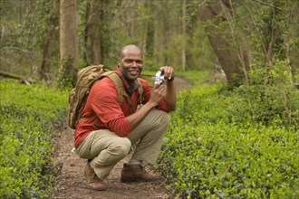 African American man crouching on path in forest holding camera