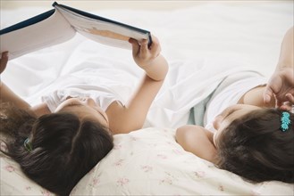 Two young Hispanic sisters reading a book in bed