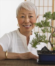Senior Asian woman smiling with bonsai tree indoors