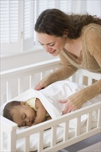 Hispanic mother tucking sleeping baby in crib