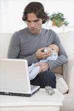 Hispanic father feeding baby and looking at laptop