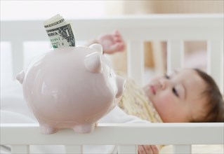 Mixed race baby girl laying in crib next to piggy bank