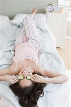 Mixed race woman placing cucumbers over eyes in bed