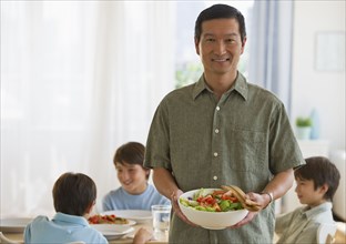 Smiling father holding salad with sons at table in background