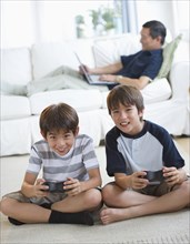Smiling twin brothers playing video game while father uses laptop on living room sofa
