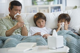 Sick father and sons blowing noses and checking temperature on living room sofa