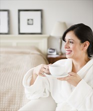 Hispanic woman in bathrobe drinking coffee in bedroom