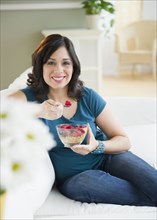Smiling Hispanic woman eating cereal on sofa