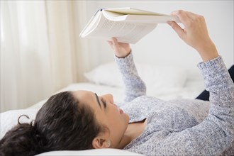 Hispanic woman reading book on bed