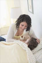 Mixed race mother tucking daughter and teddy bear into bed