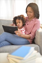 Black mother and daughter using digital tablet on sofa