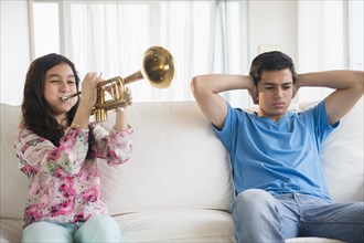Hispanic brother covering his ears as sister practices trumpet in living room