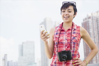 Mixed race woman with camera and cell phone smiling near city skyline