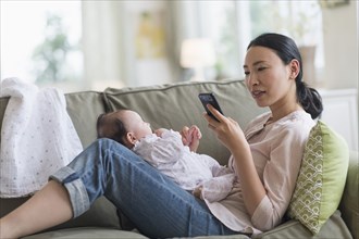Asian mother using cell phone and holding baby in living room