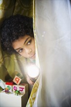 Mixed race boy with flashlight peering out of blanket fort