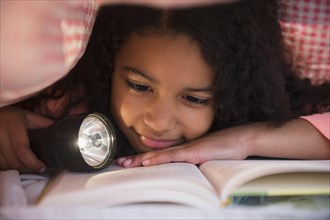 Mixed race girl reading book with flashlight under blanket