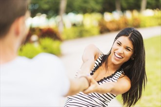 Smiling Hispanic woman pulling boyfriend in park