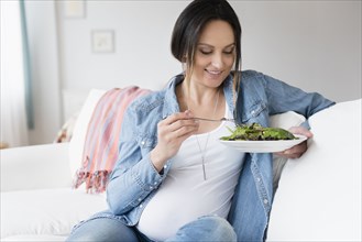 Pregnant Caucasian woman eating salad on sofa