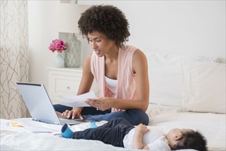 Mother paying bills on laptop with baby son on bed