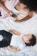 Mother drinking coffee while watching baby son sleeping on bed