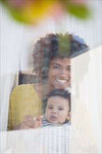Mother holding baby son behind window curtain