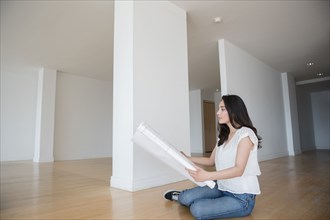 Hispanic woman sitting on floor of empty apartment holding blueprint
