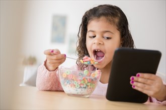 Mixed Race girl eating cereal and using digital tablet
