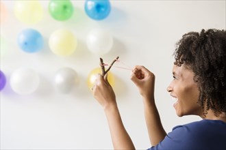 Black woman aiming slingshot at balloons on wall