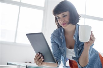 Hispanic woman drinking coffee and reading digital tablet