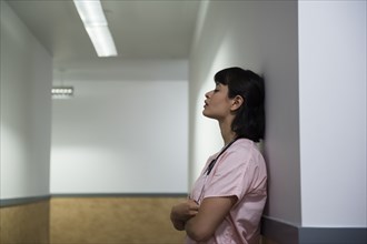 Tired Hispanic nurse leaning on wall in hospital corridor
