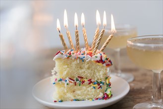 Candles burning on slice of cake with sprinkles near champagne