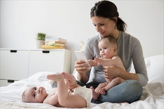 Caucasian mother with twin baby daughters on bed texting on cell phone