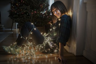 Mixed Race woman sitting on floor wrapped in string lights near Christmas tree