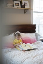 Caucasian baby girl sitting on bed reading book