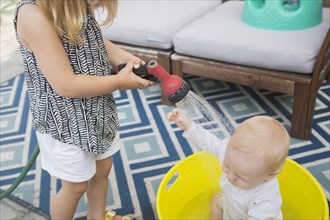 Caucasian girl spraying baby brother in bucket with garden hose