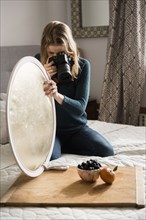 Woman kneeling on bed holding reflector photographing fruit