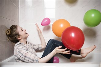 Laughing woman sitting in bathtub playing with multicolor balloons