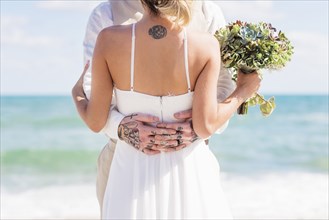 Caucasian bride and groom with tattoos hugging on beach