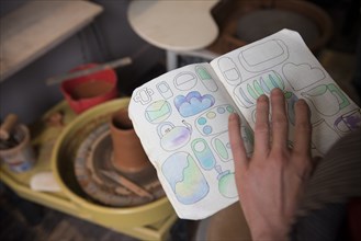 Hand of Caucasian woman examining drawings in book near pottery wheel