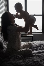 Caucasian mother sitting on bed lifting baby son